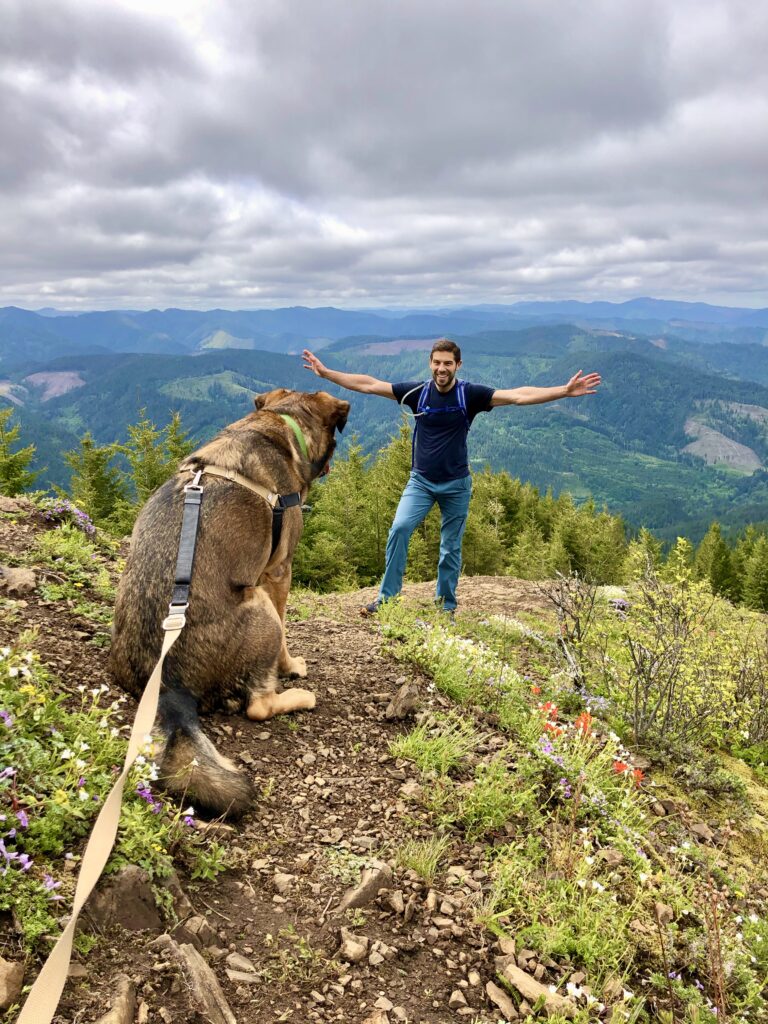 seated dog in the foreground with a man standing in the background with his arms outstreached